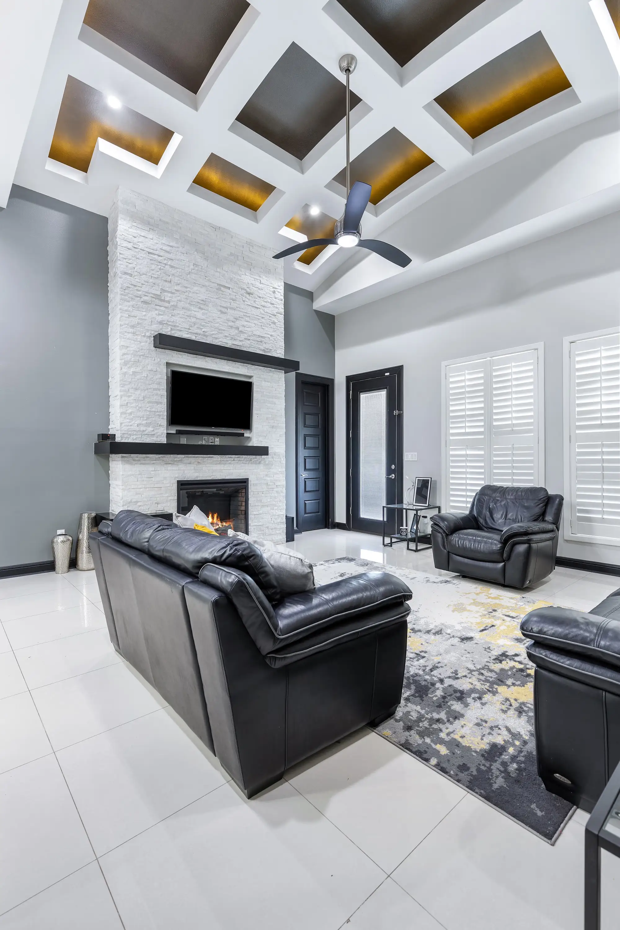 Living room under a lit coffered ceiling with a stacked-stone fireplace and media wall